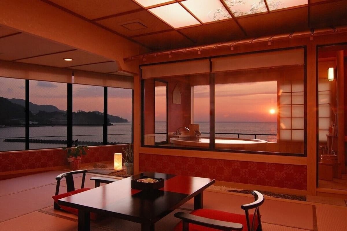 A room with a large window overlooking a body of water and mountains, a table with chairs, and a small sitting area with a bench.