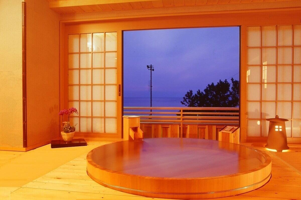 A traditional Japanese onsen with a wooden interior, a tatami mat floor, a wooden bench, and a view of the sea.