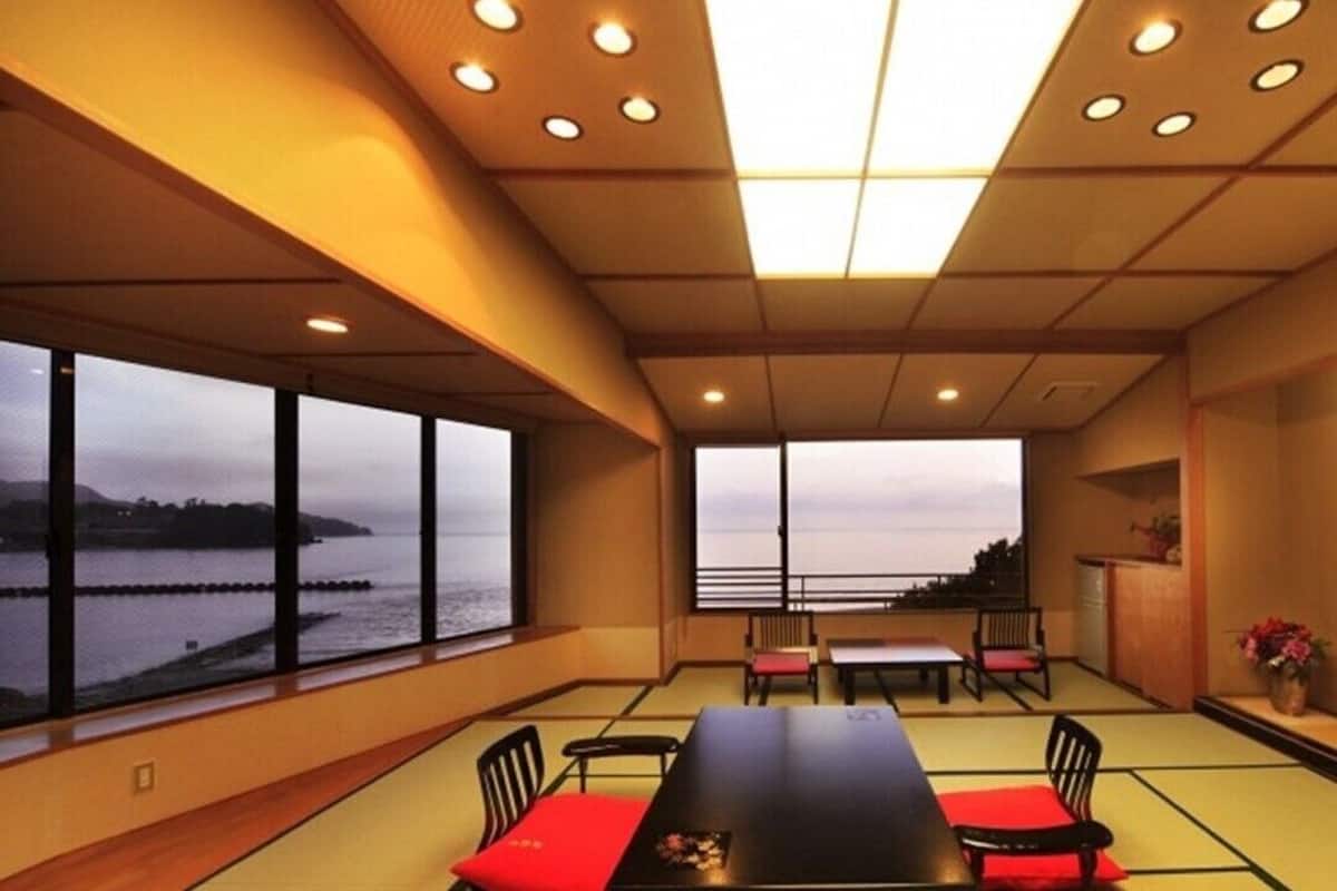 A room with tatami mats, a low table, chairs, a large window with a view of water, and recessed lighting.