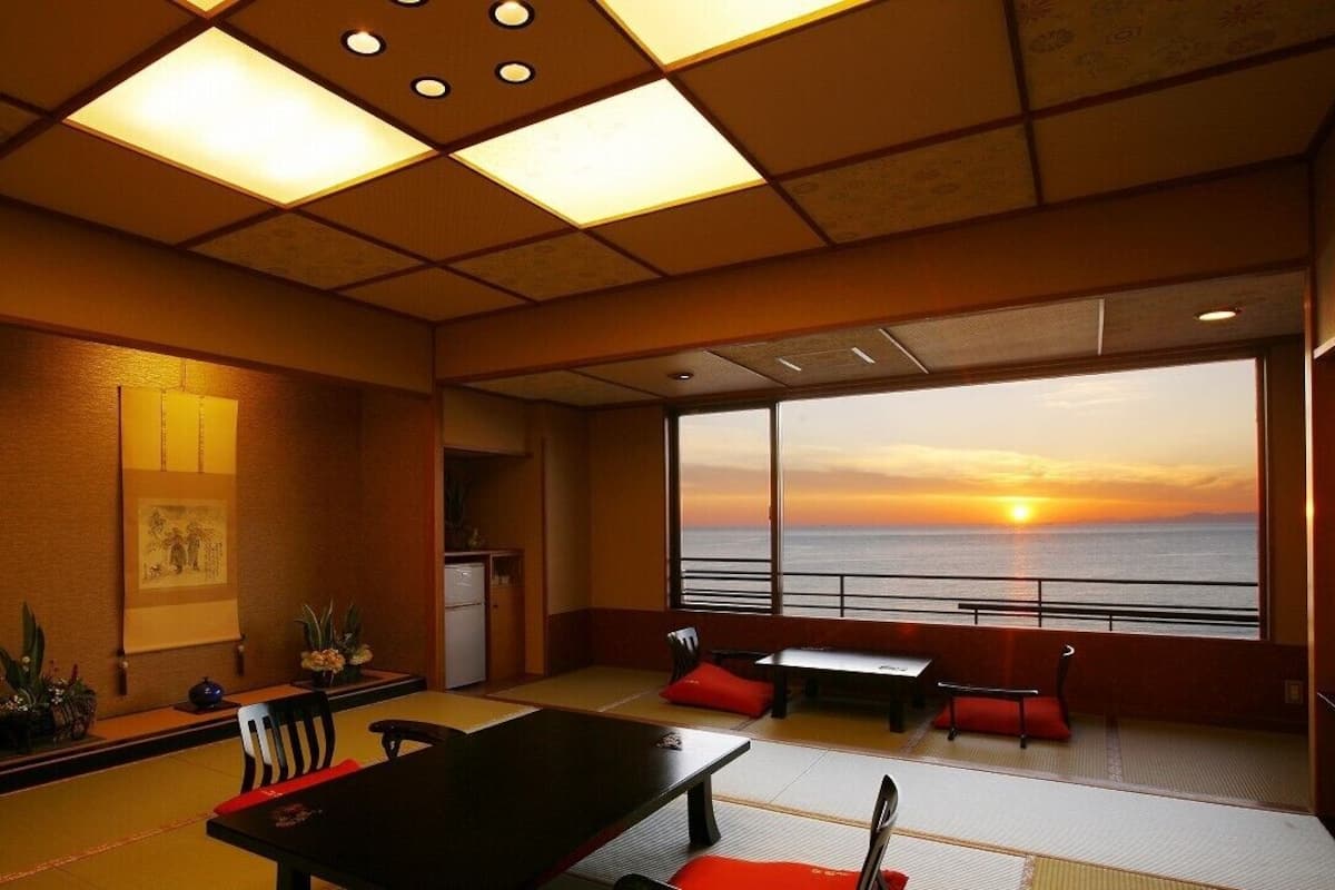 A room with tatami flooring, a low table, chairs, a small refrigerator, and a large window with a sunset view.