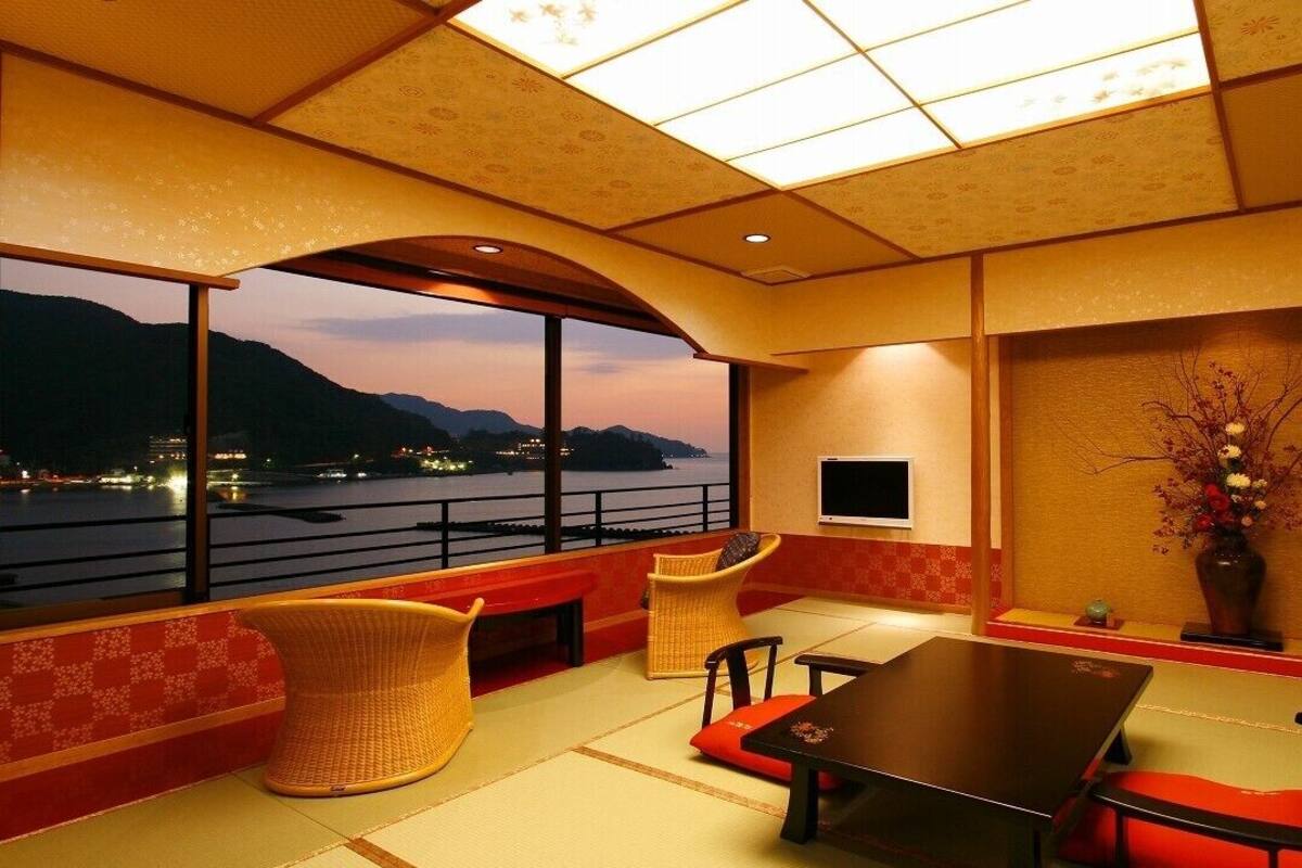 A room with a large window overlooking a body of water, a dining area with a table and chairs, and a wall-mounted television.