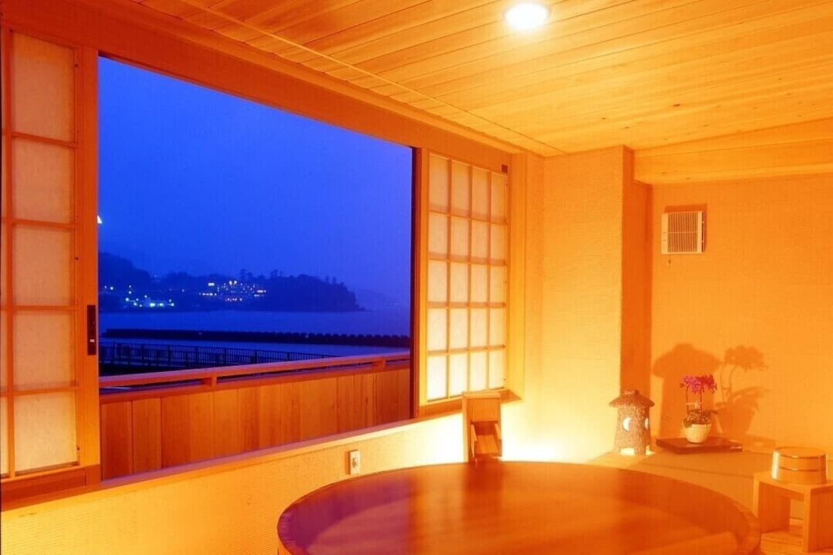 A room with a large window, wooden flooring, a round table, and a small shelf with decorative items.