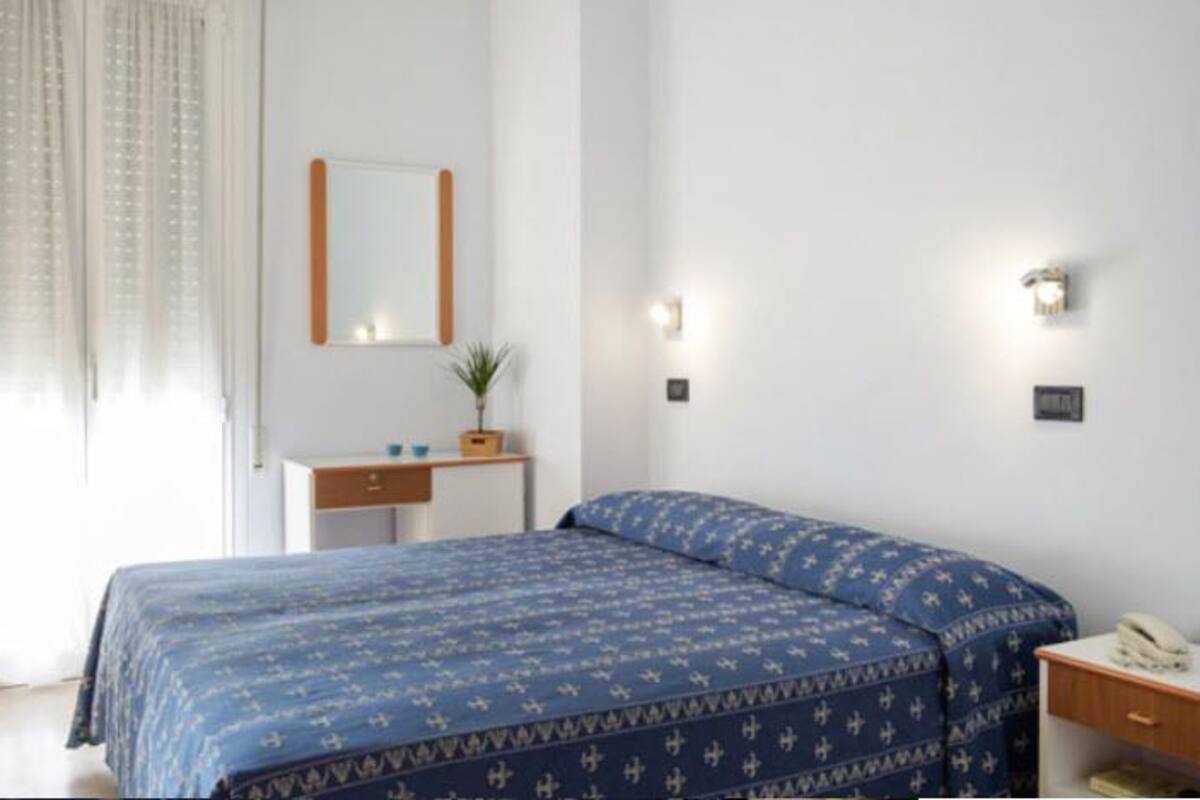 A bedroom with a bed, a bedside table, a mirror, and a window with curtains.