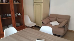 Une pièce avec un canapé beige, une chaise blanche, une table en bois et un buffet contenant de la vaisselle.