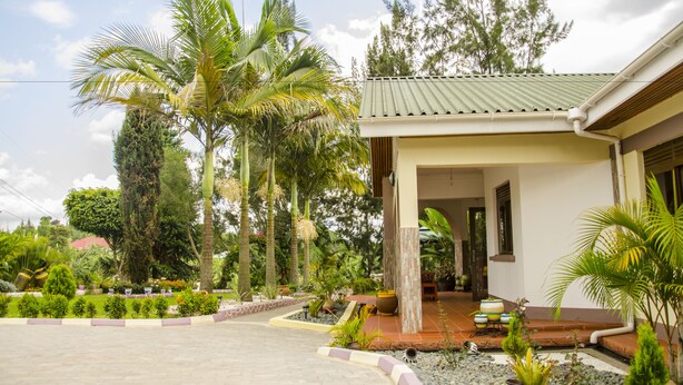 A house with a covered porch, surrounded by a garden with various plants and trees. - Sheevy's Bed N Breakfast (Fort Portal)