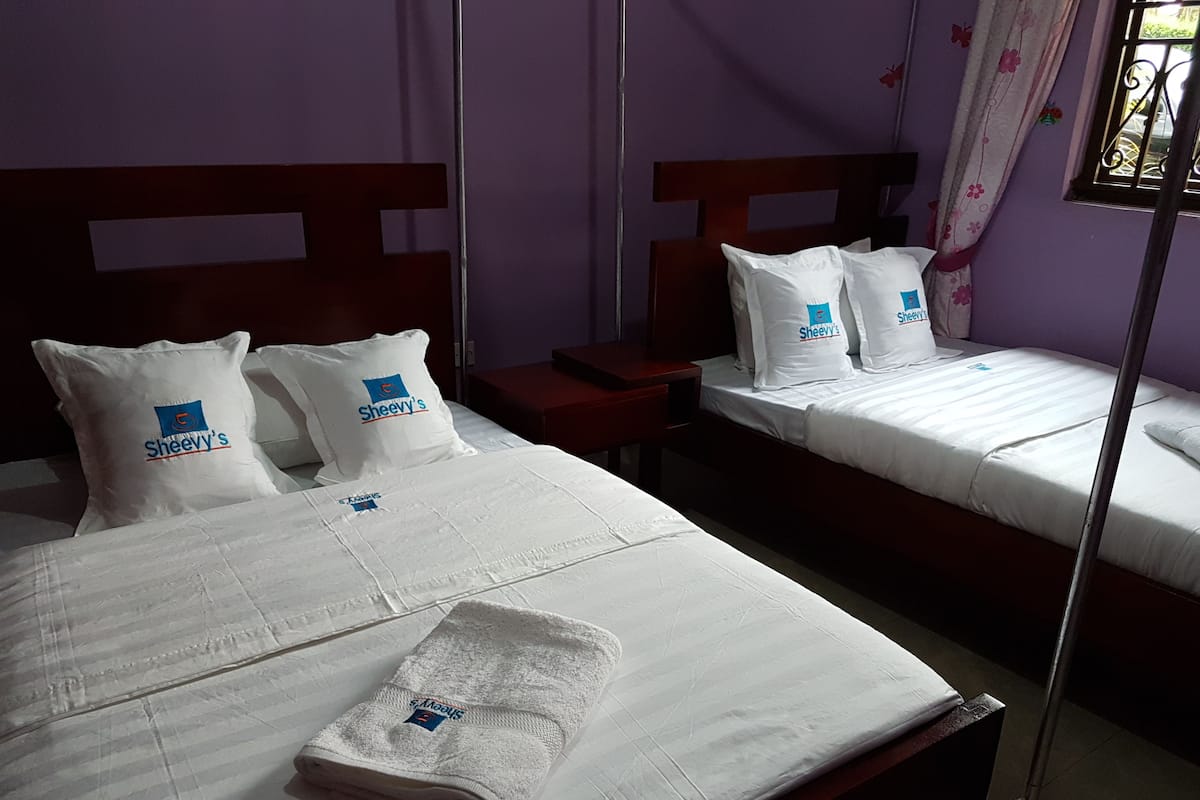A hotel room with two beds, each with white bedding and Sheevy's logo pillows. There is a folded towel on the bed in the foreground.