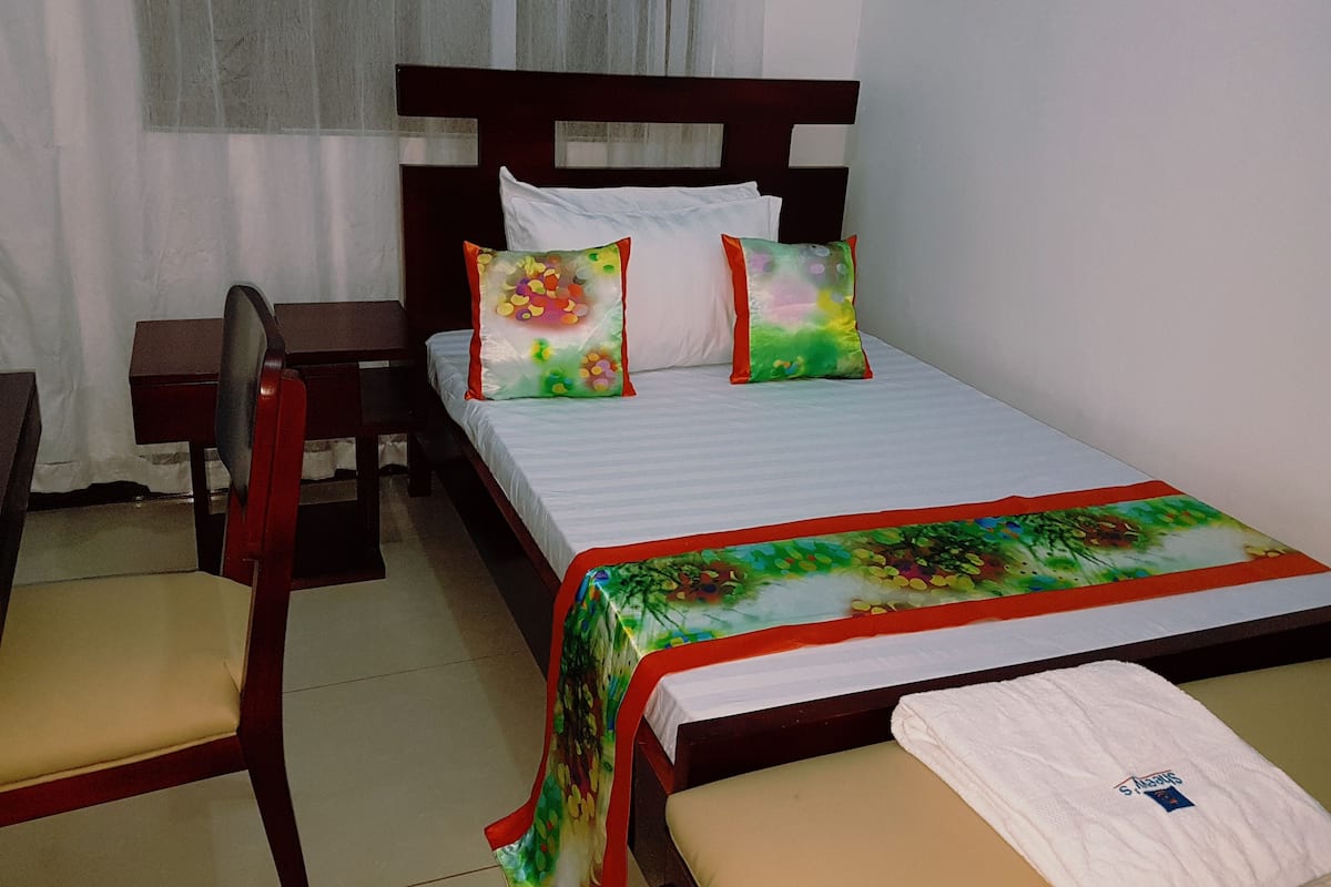 A single bed with a floral bedspread, two pillows, a wooden headboard, a small bedside table, and a chair.