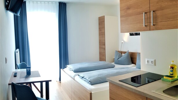 Habitación de hotel pequeña con cama, escritorio, silla, televisor montado en la pared y armarios de madera.