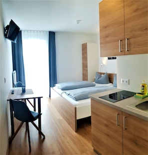 Habitación de hotel pequeña con cama, escritorio, silla, televisor montado en la pared y armarios de madera.