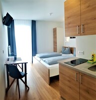 Quarto de hotel pequeno com uma cama, uma mesa, uma cadeira, uma TV fixada na parede e armários de madeira.