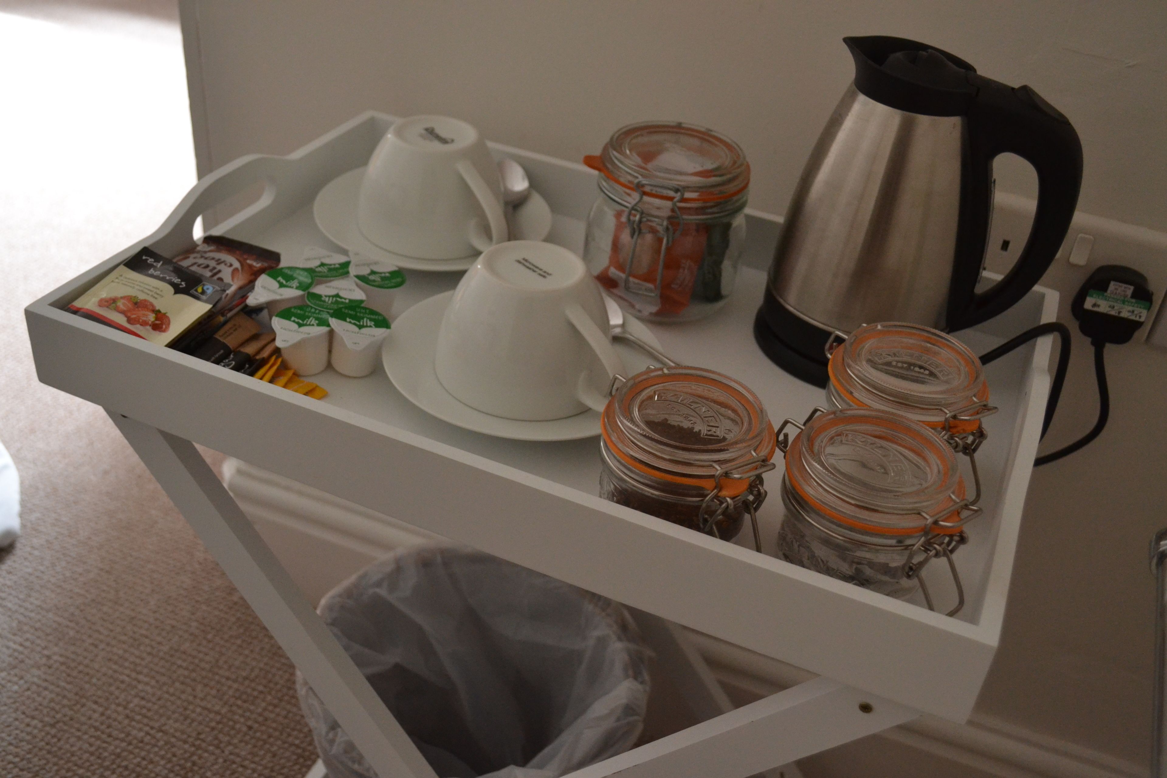 A white tray table with a coffee maker, cups, jars, and a trash bin.