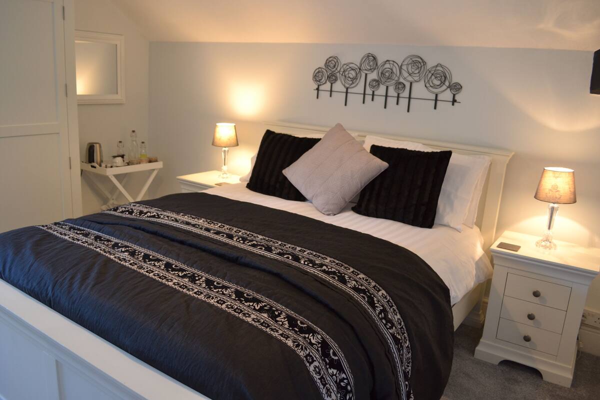 Una cama bien hecha con colcha de cuadros blanco y negro, dos mesitas de noche con lámparas, un adorno en la pared y un mueble auxiliar blanco con cajones.