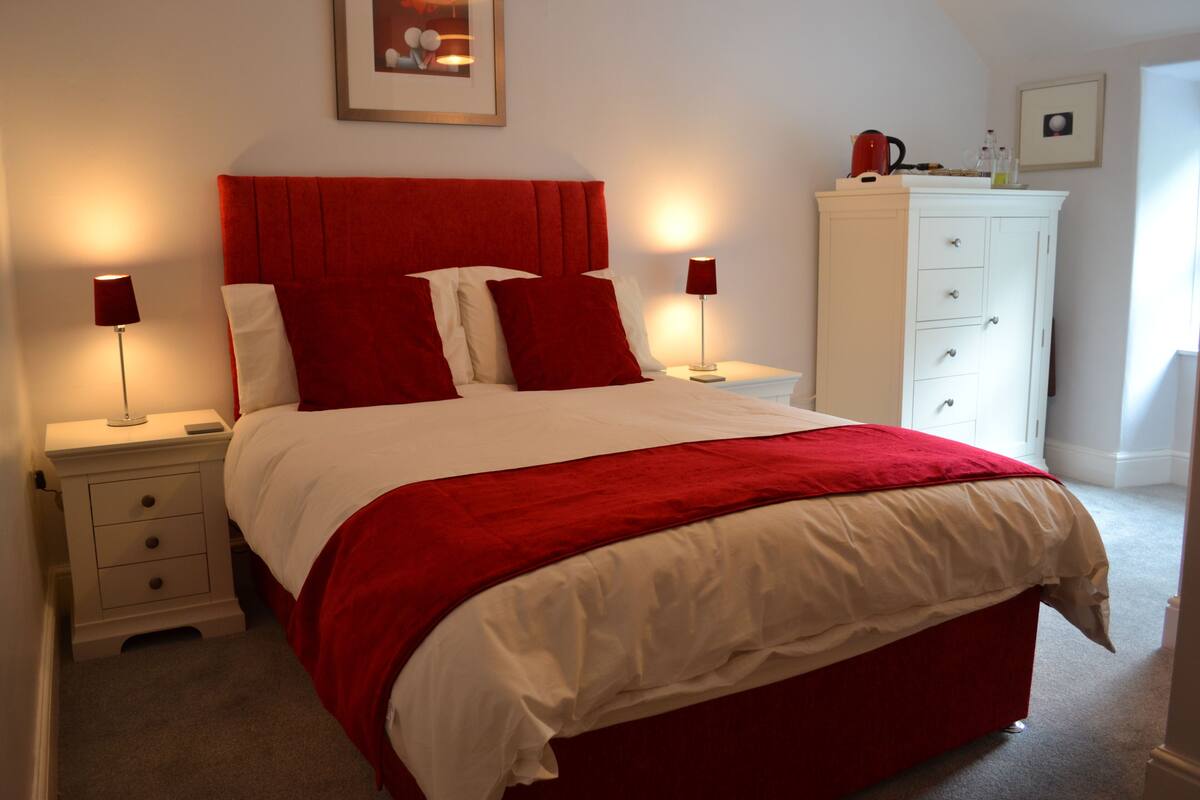 Una cama bien hecha con ropa de cama roja y blanca, dos mesitas de noche con lámparas, un aparador blanco y un cuadro en la pared.