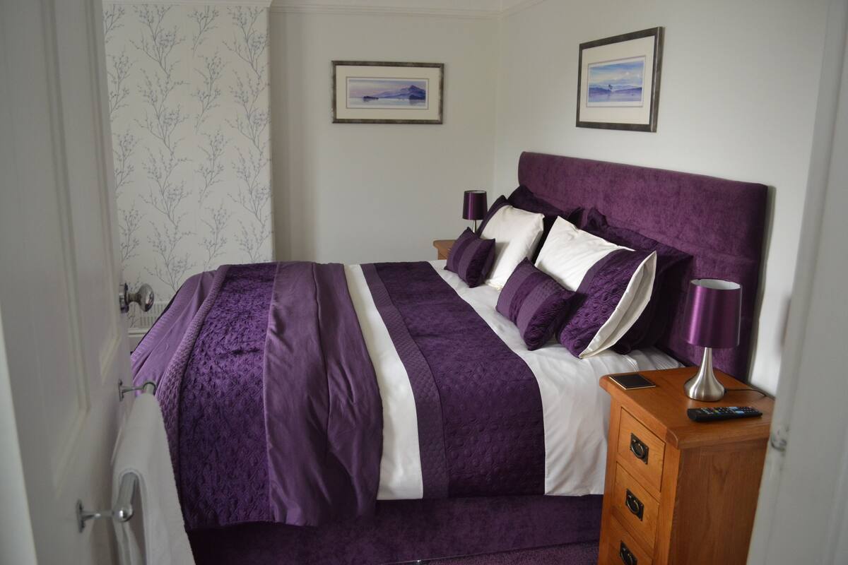 Una cama bien hecha con cubrecama morado y blanco, una mesita de noche de madera con una lámpara y dos cuadros en la pared.