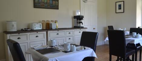 A dining area with a white table set for four, a coffee maker, and a painting on the wall.