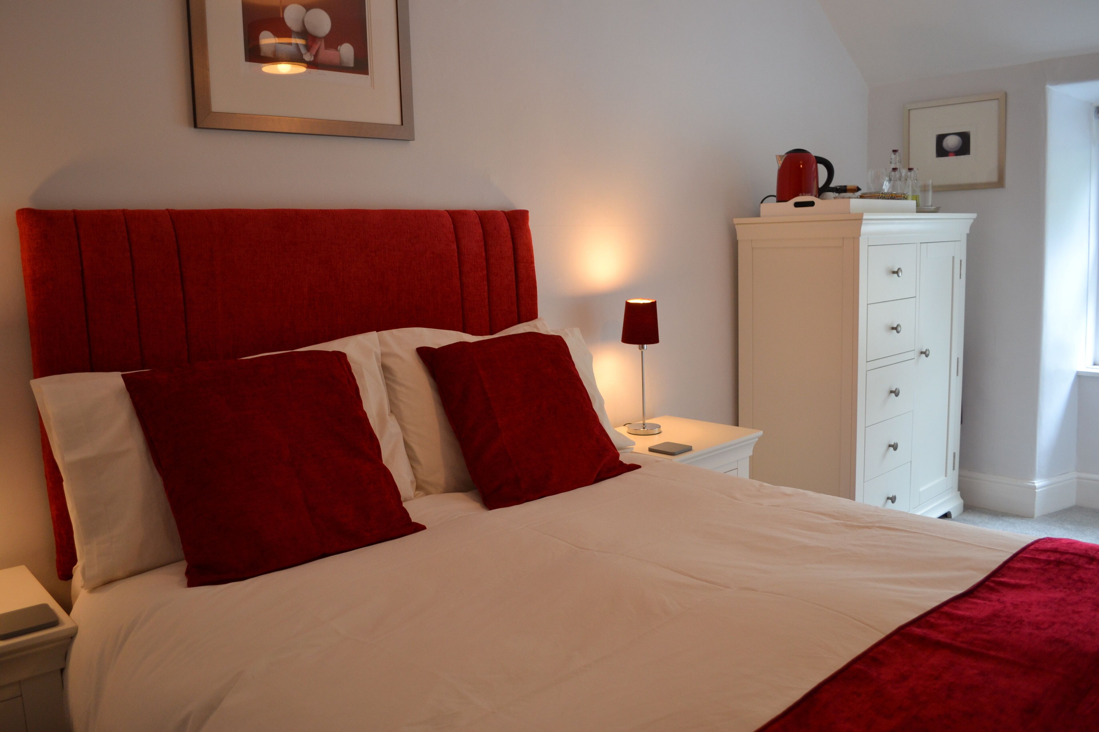 Un lit bien fait, avec une literie rouge et blanche, une table de chevet avec une lampe et une commode blanche avec des tiroirs.