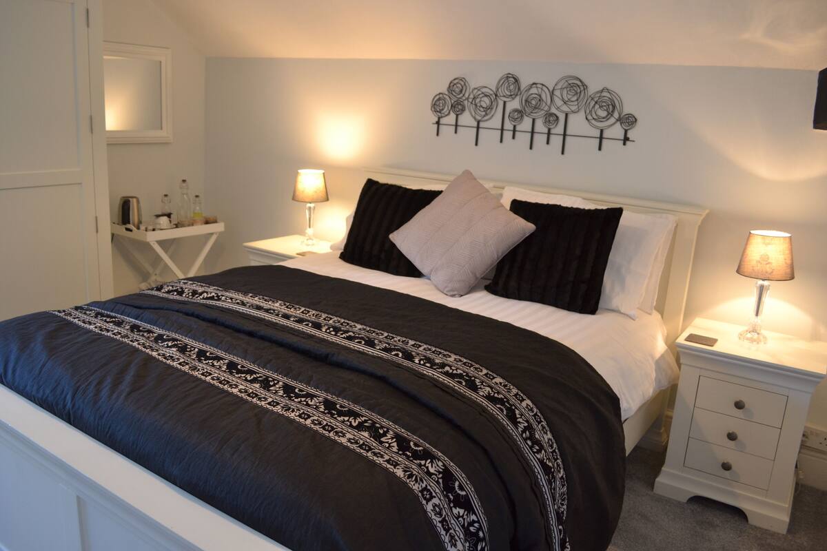Una cama bien hecha con colcha blanco y negro estampada, dos lámparas de noche, una mesita de noche con lámpara y una decoración en la pared sobre la cama.