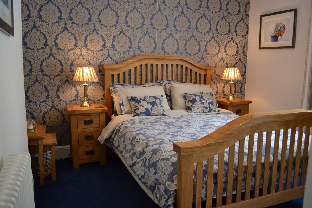 Un dormitorio con cama de madera, ropa de cama estampada con motivos florales, dos lámparas de noche y un papel tapiz azul y blanco con diseño damasco.