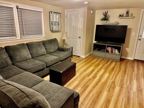Una sala de estar con un sofá seccional, un televisor sobre un mueble, una lámpara y una estantería.