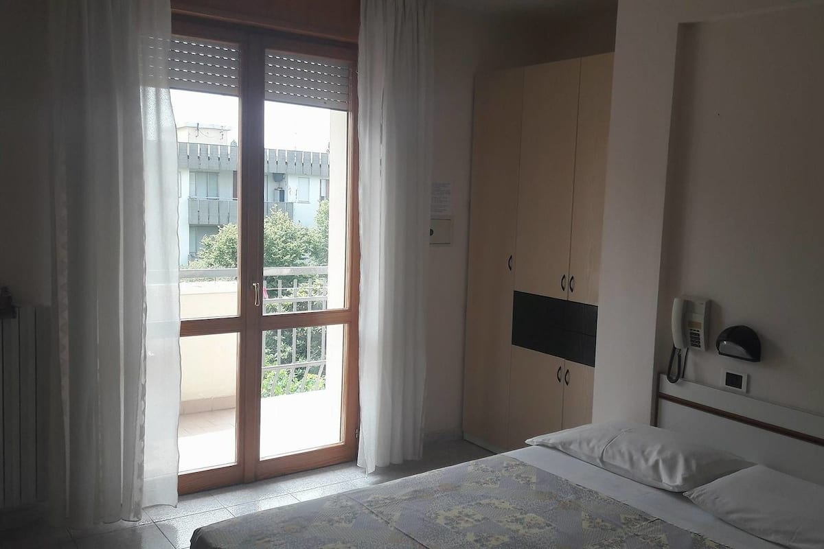 Habitación de hotel con cama, armario, escritorio con teléfono y vista al exterior.
