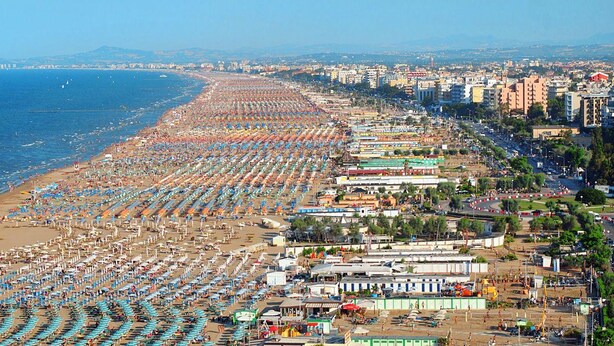 Un paysage urbain côtier avec une longue plage bordée de rangées de parasols et de personnes se prélassant au soleil.