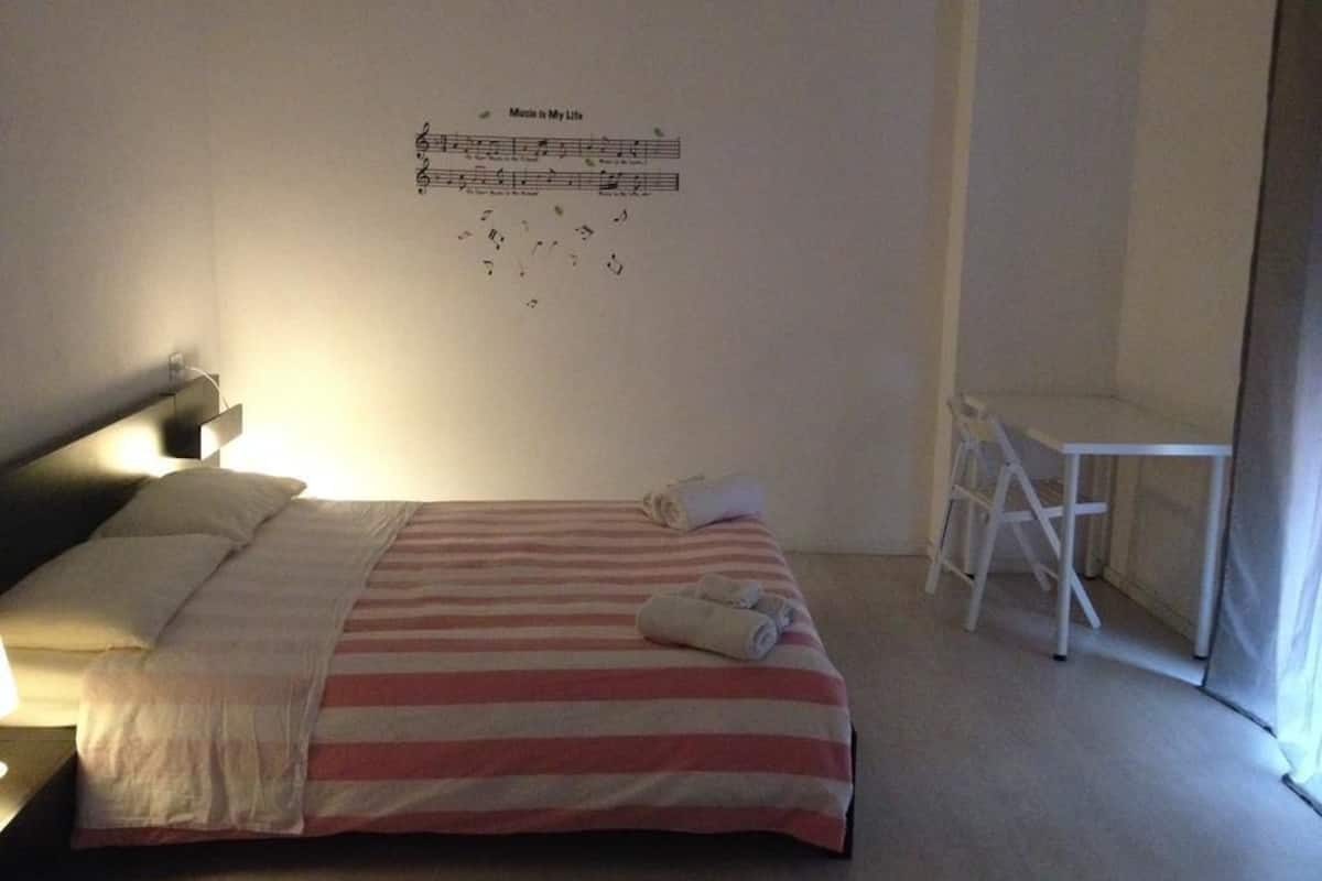 Una cama bien hecha con ropa de cama a rayas, una mesita pequeña y una silla en una habitación sencilla con una partitura musical colgada en la pared.