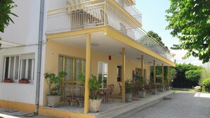 Un bâtiment à deux étages, doté d’une façade jaune, de balcons avec des garde-corps en métal et d’un espace extérieur aménagé avec des plantes en pot.
