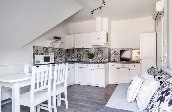 A compact kitchen with white cabinets, a microwave, and a TV. There is a dining area with a white table and chairs.