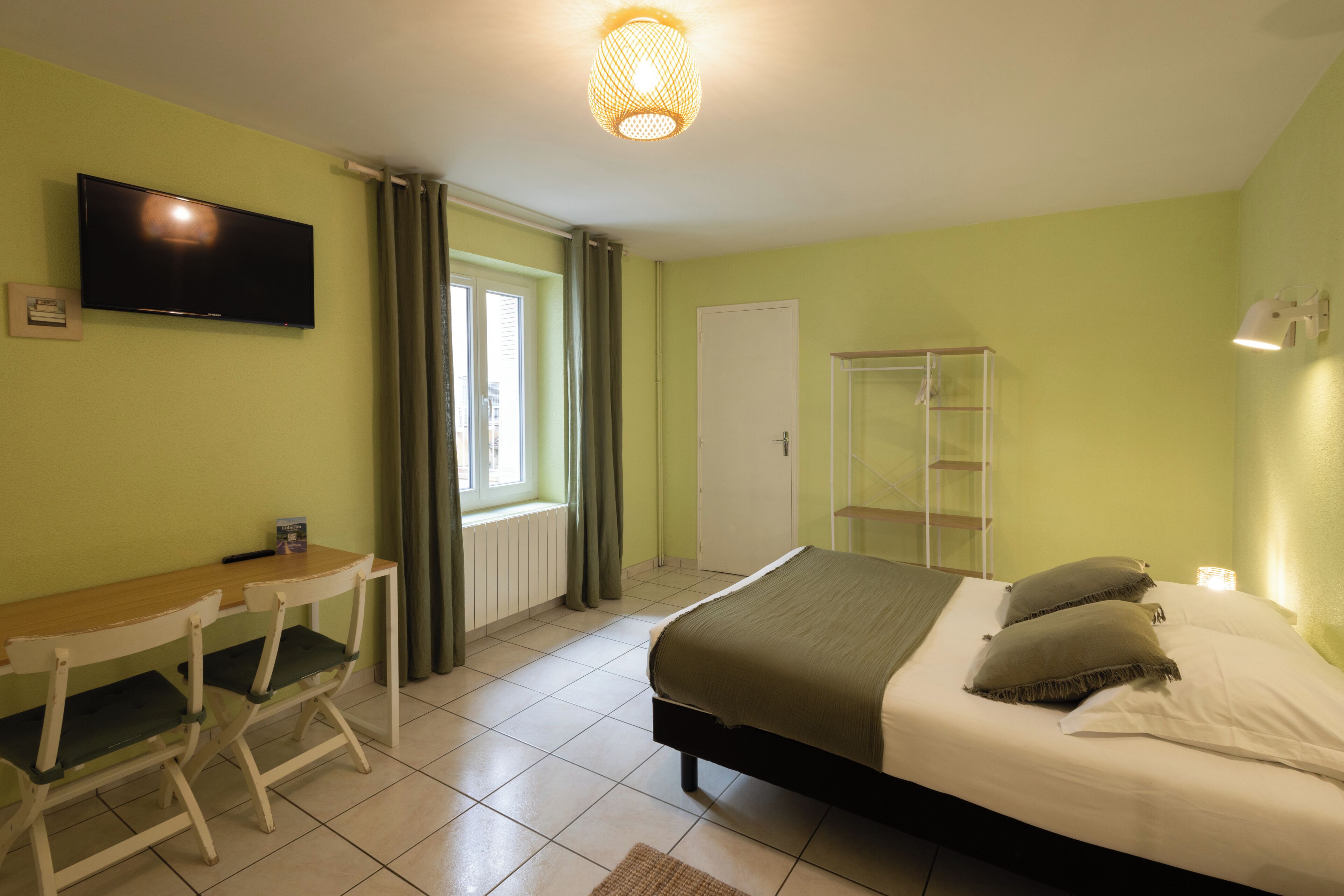 Ein Zimmer mit einem Bett, einem Schreibtisch, einem Fernseher, einem Fenster mit Vorhängen und einer Tür.