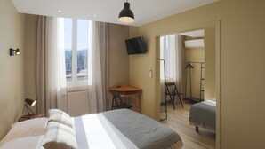 A hotel room with a bed, a desk, a chair, a TV mounted on the wall, and a view of the city through the window. - L'Aptois Hotel (Apt)