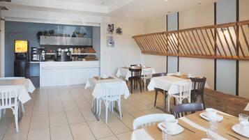 A modern dining area with white tables and chairs, a kitchen in the background, and a wooden slatted ceiling design.