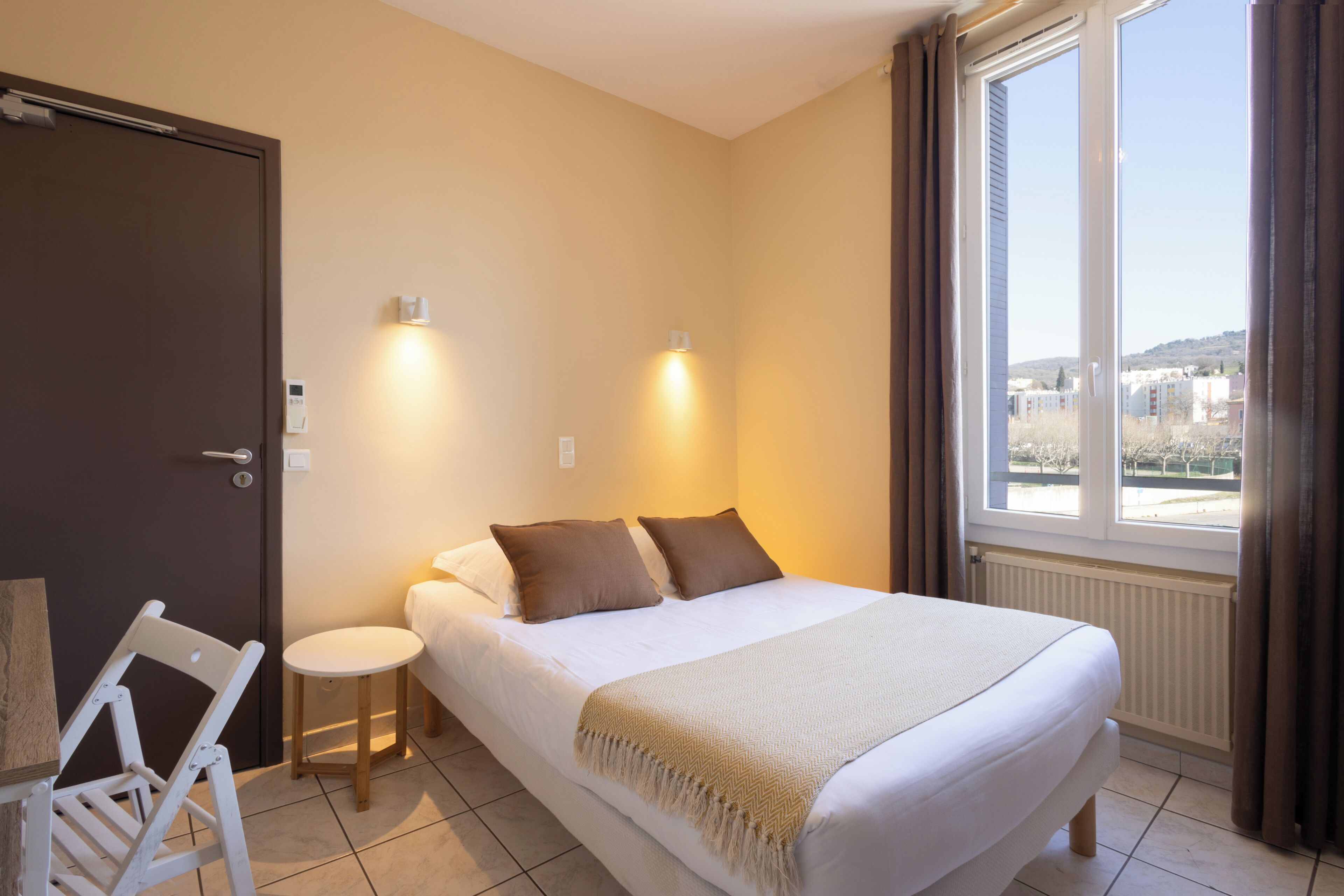 A bedroom with a bed, bedside lamps, a chair, a small table, and a window with a view.