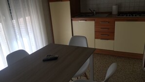 Un coin repas avec une table en bois, des chaises blanches, une télécommande et un espace cuisine comprenant un évier et des placards.