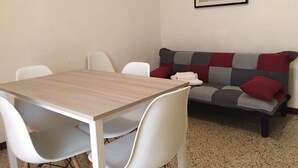 Une pièce avec une table en bois et des chaises blanches, un canapé gris et rouge avec un plaid, et un tableau encadré au mur.