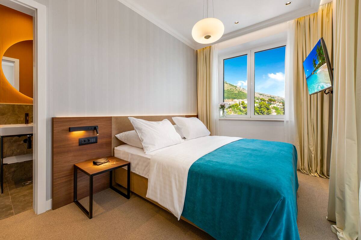 A hotel room with a bed, a small table, a TV mounted on the wall, and a window with a view of a landscape.