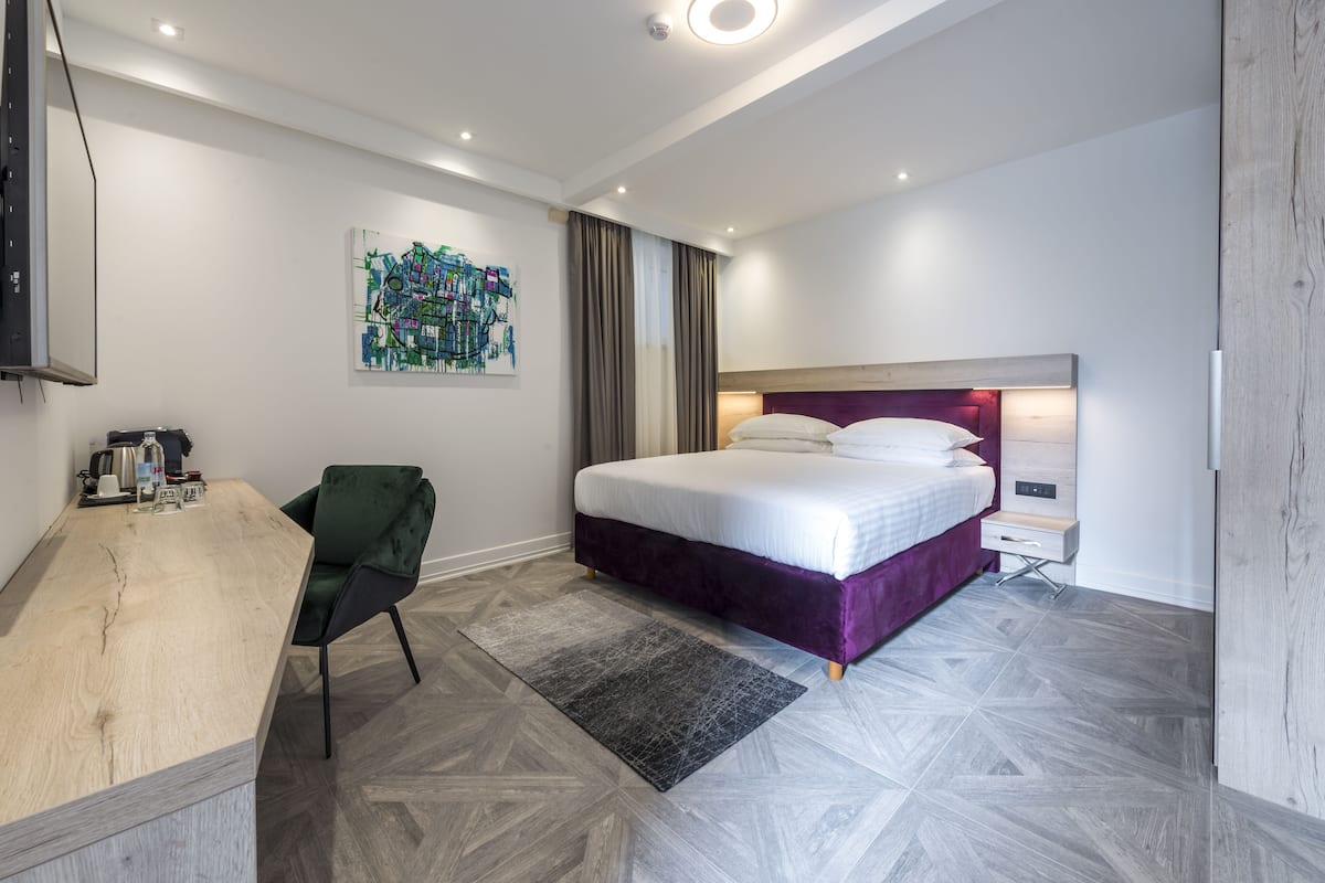 A modern hotel room with a bed, a desk, a chair, and a painting on the wall.
