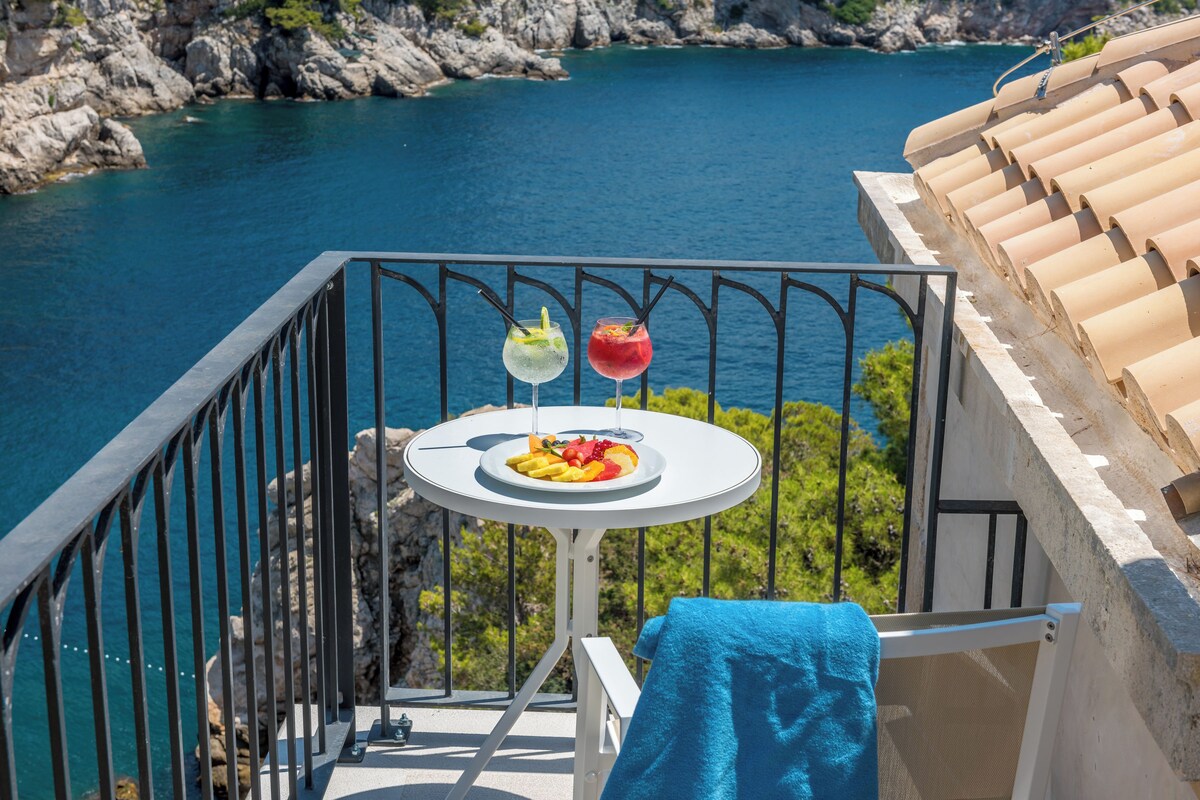 Un balcon avec une table dressée pour deux personnes, offrant une vue sur une zone côtière avec une eau d’un bleu limpide et un paysage rocheux.