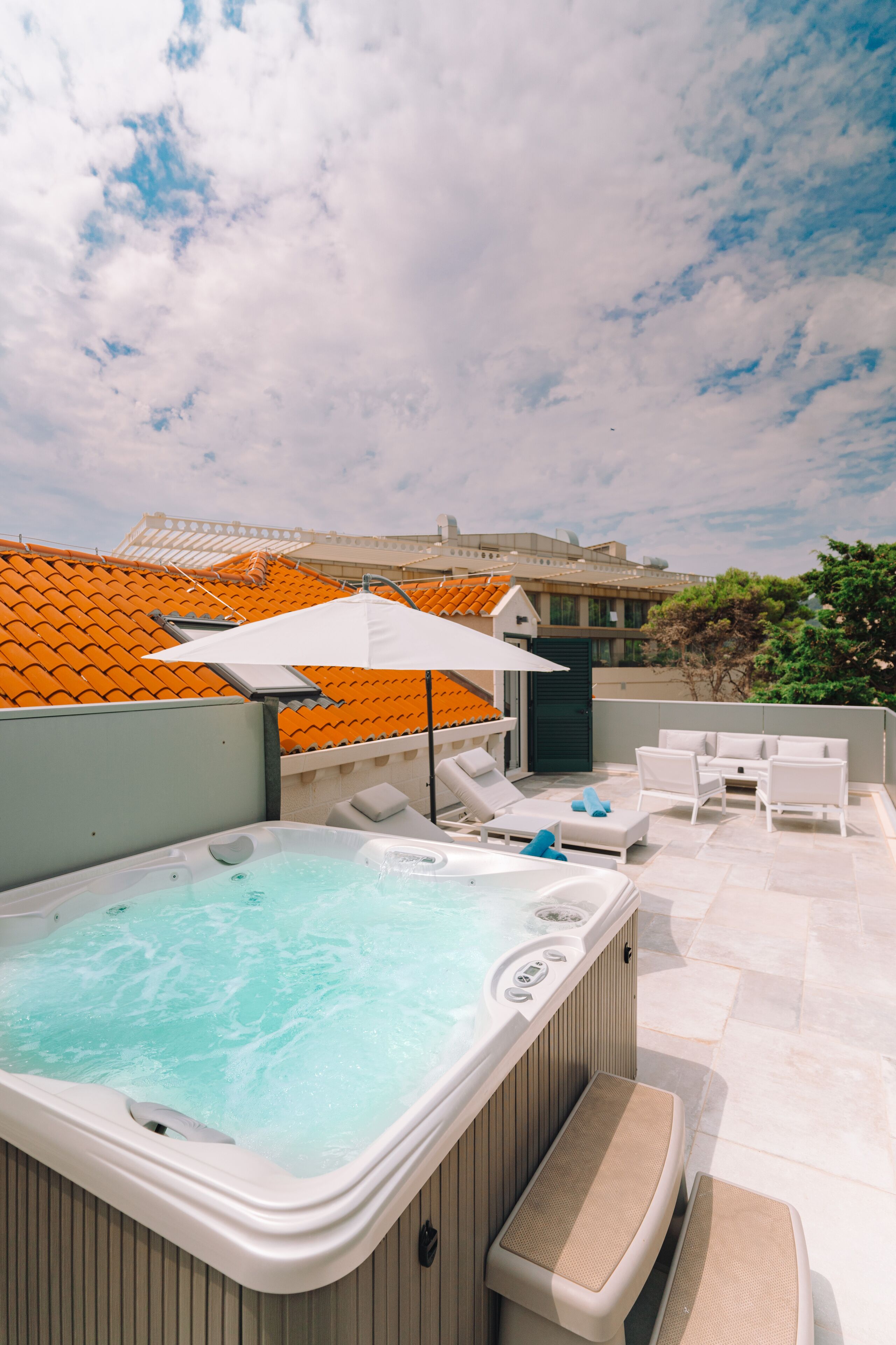 A rooftop hot tub with lounge chairs and an umbrella, surrounded by orange-tiled roofs and greenery.