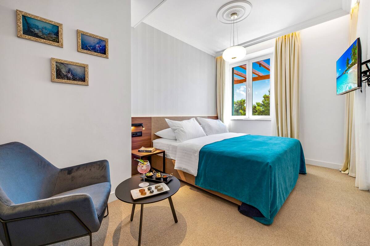 A hotel room with a bed, a chair, a small table with snacks, and three framed pictures on the wall.
