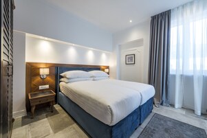 Quarto de hotel com uma cama grande, uma mesa de cabeceira com abajur e vista para o exterior pela janela.