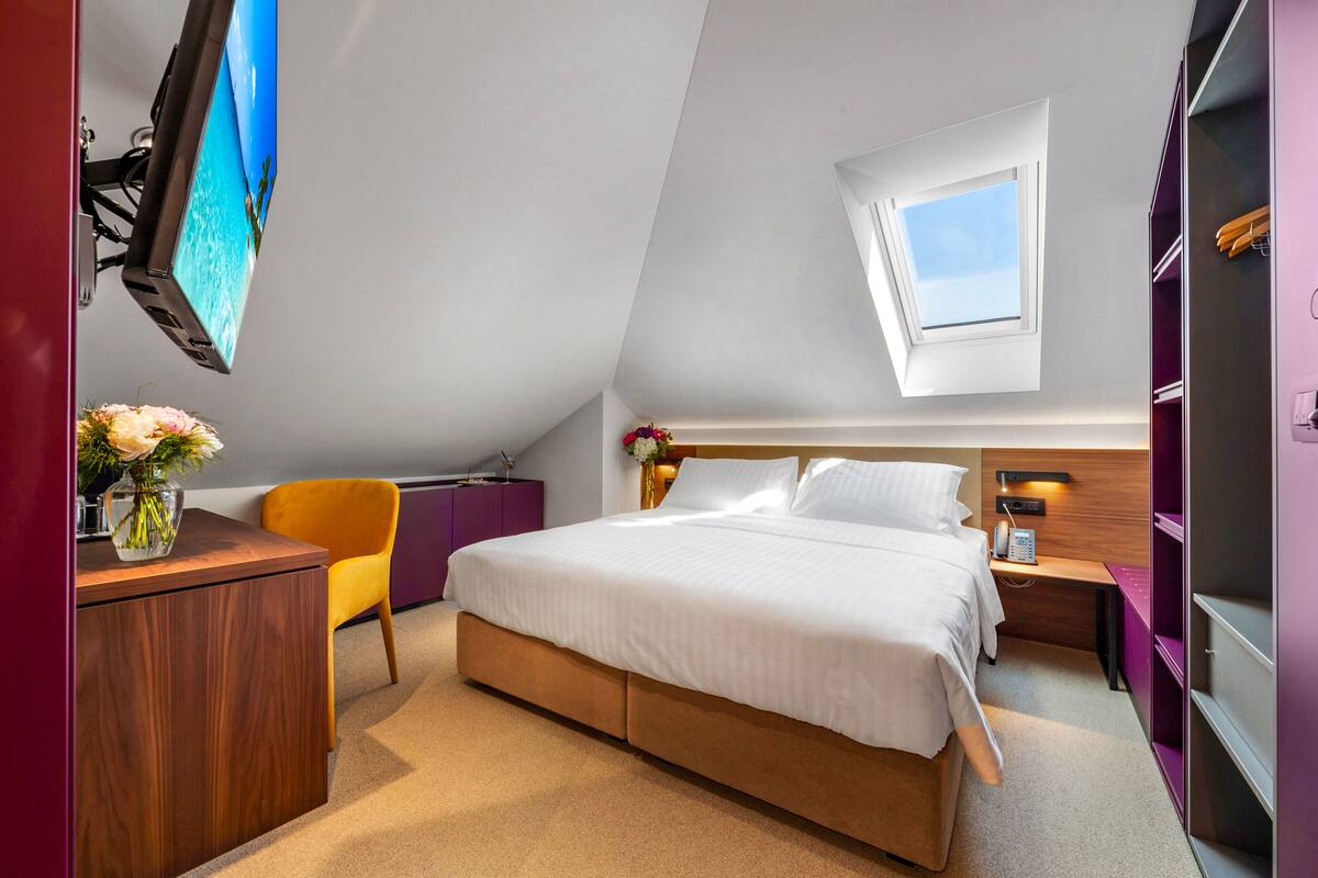 A bedroom with a bed, a desk, a chair, a nightstand, a TV, and a window.
