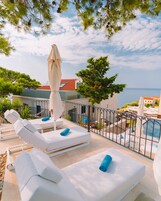 Terraço no topo com cadeiras de lounge brancas e piscina, com vista para uma área costeira com edifícios e árvores.