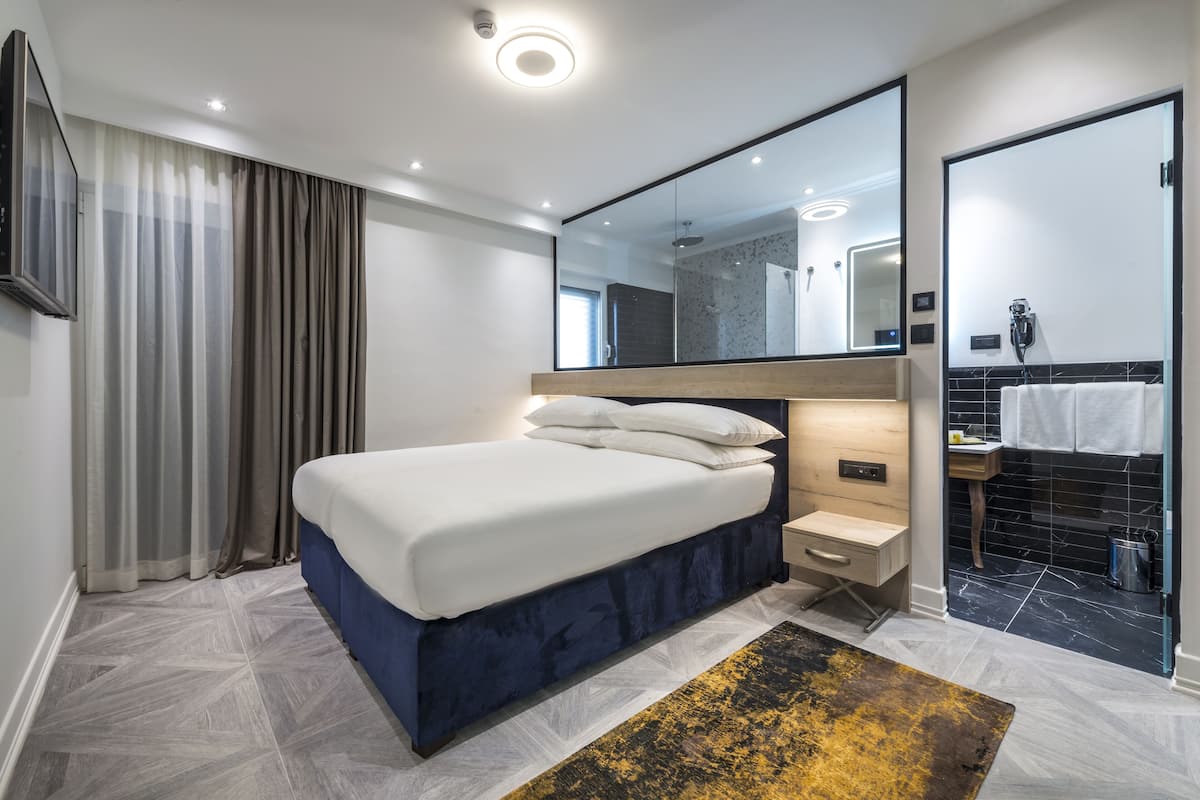 A modern bedroom with a large bed, bedside tables, a mirror, and a bathroom with a shower and sink.