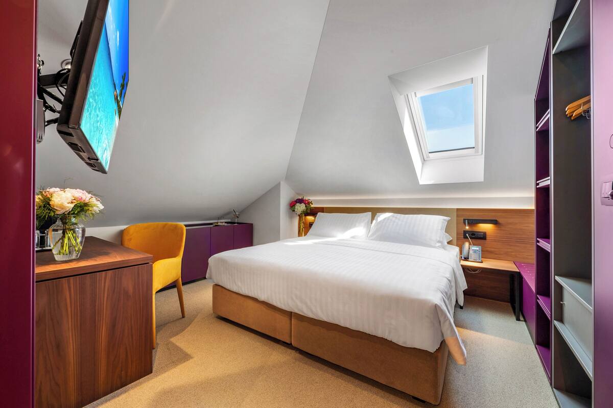 A bedroom with a bed, a desk, a chair, a nightstand, a TV, and a window.