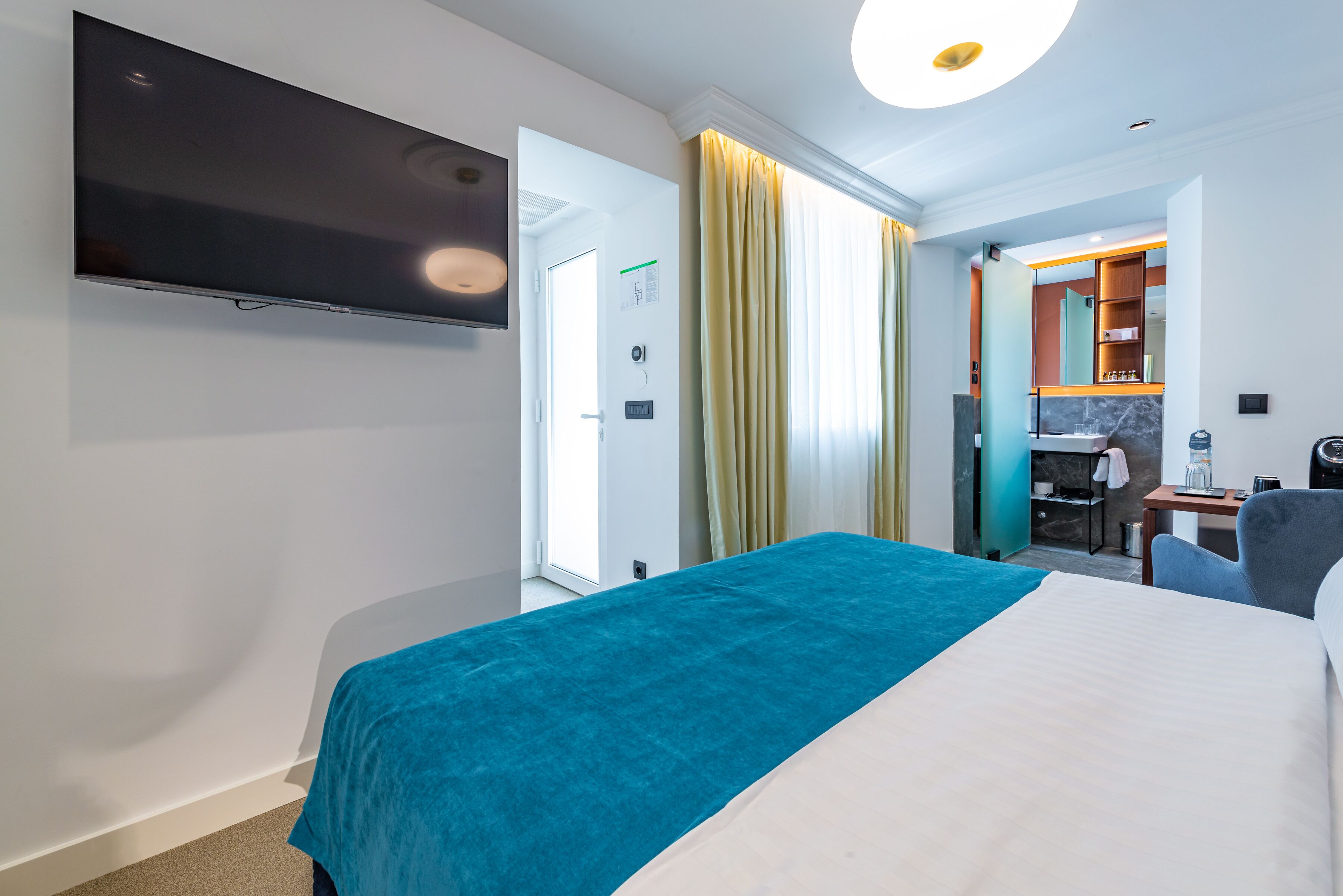 Quarto de hotel com cama, televisão, secretária e casa de banho visível por uma porta aberta.