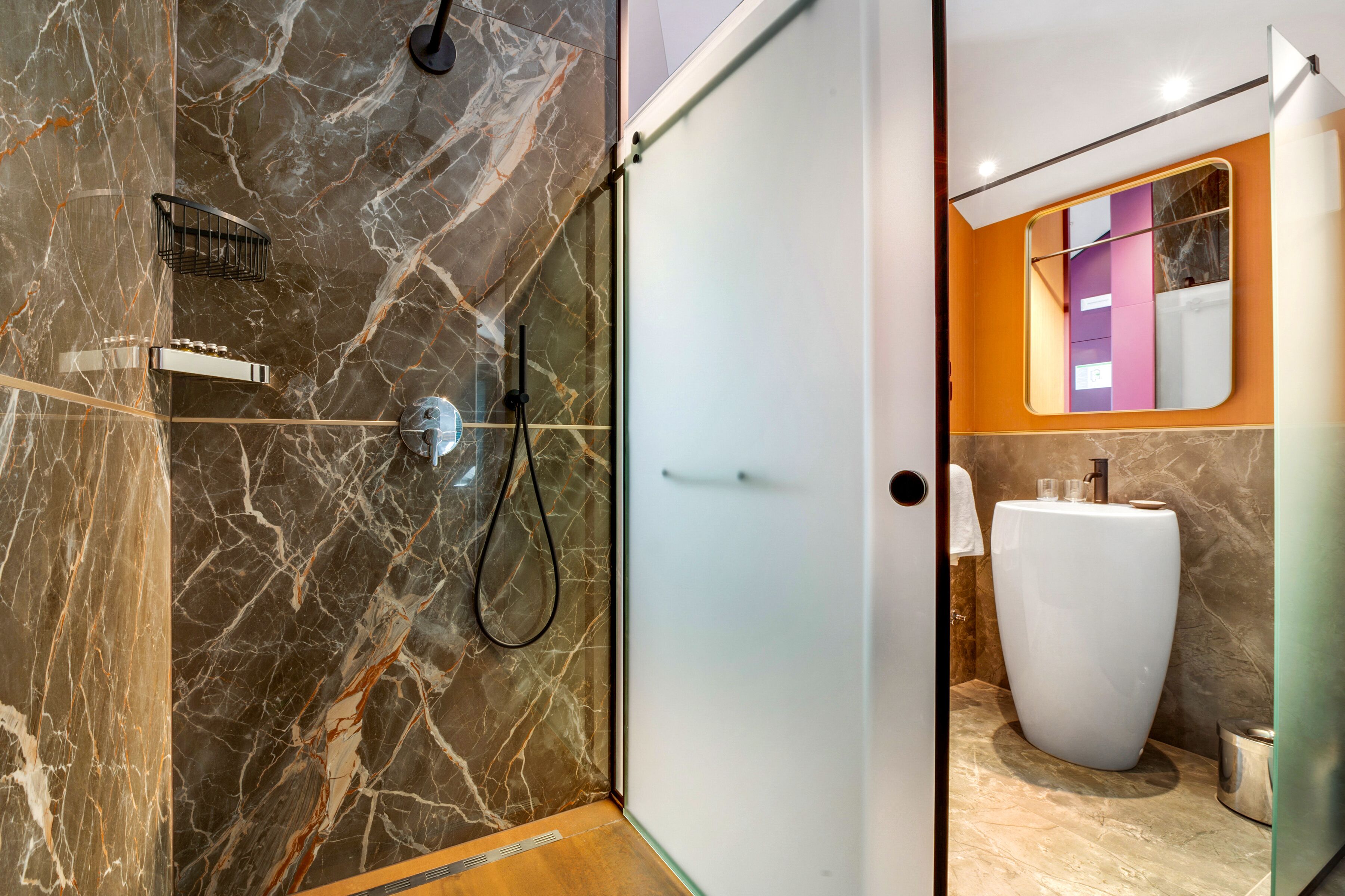 A modern bathroom with a marble wall, a glass shower enclosure, a white sink, and a mirror with a colorful frame.