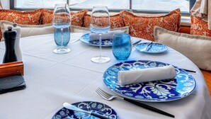 Mesa de jantar posta para dois, com pratos azuis e brancos estampados, copos e uma garrafa de água. A mesa está coberta por uma toalha branca, e existem almofadas laranja e brancas estampadas no sofá. É visível uma grande janela com vista para o oceano.