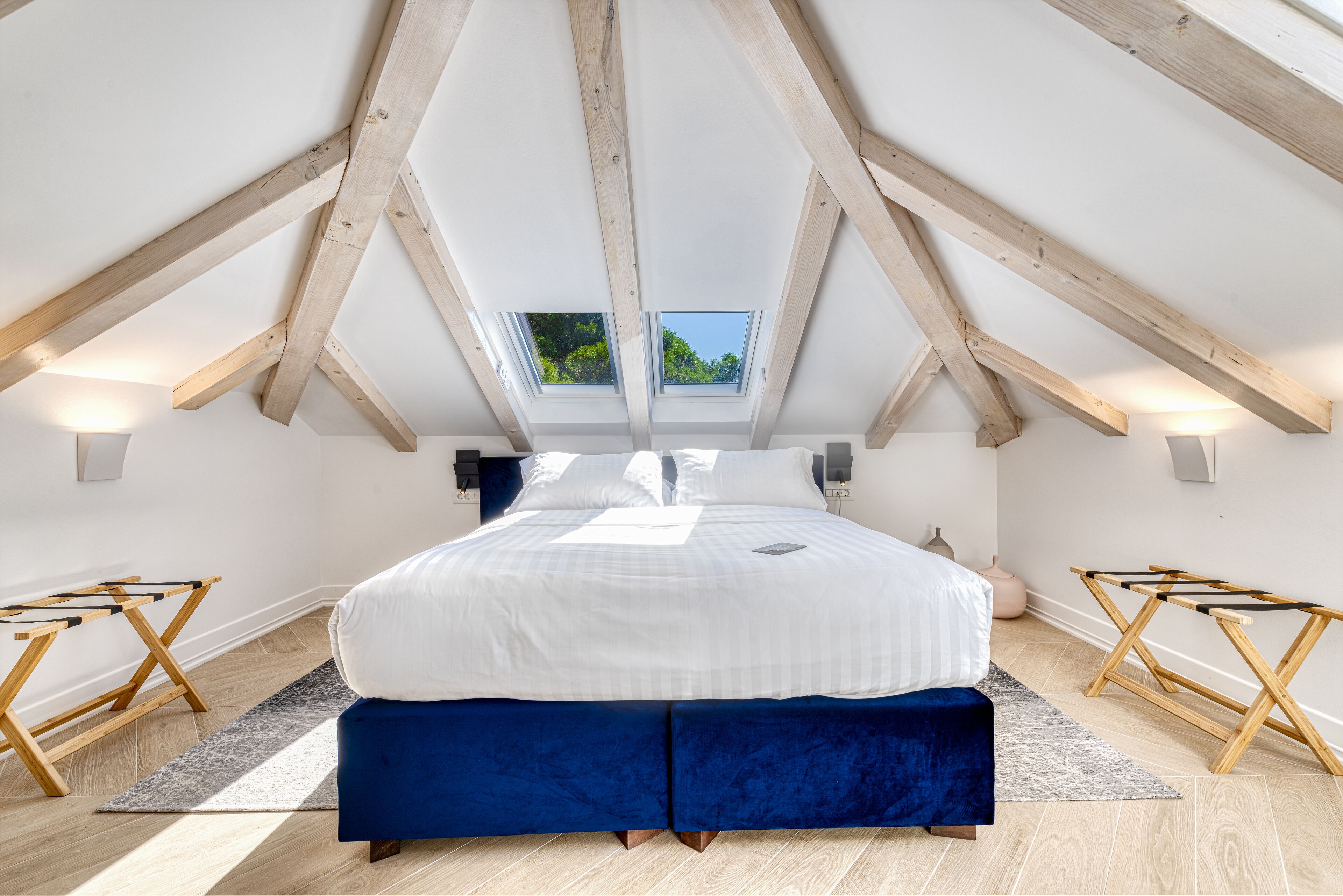 A bedroom with a sloped ceiling, skylight, wooden beams, a large bed, bedside tables, and a view of greenery outside.
