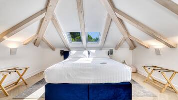 A bedroom with a sloped ceiling, skylight, wooden beams, a large bed, bedside tables, and a view of greenery outside.