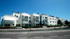 A row of modern, multi-story residential buildings with balconies, a paved road in the foreground, and a clear blue sky. - Libeccio (Jesolo)